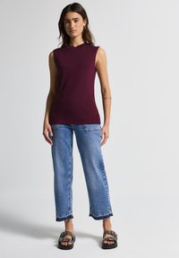 Top sin mangas burdeos con un cuello alto, combinado con jeans de pierna ancha en un tono azul claro que tienen dobladillos deshilachados; modelo con sandalias negras.