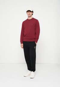 Rode crewnecktrui gecombineerd met zwarte joggingbroek met een decoratief patroon, gedragen met een witte pet en witte sneakers.