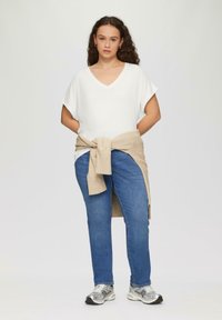 Biała koszulka z dekoltem w kształcie v, niebieskie dżinsy i srebrne sneakersy. Beżowy sweter związany w talii. Gładka tekstura, luźny krój i swobodny design.