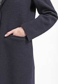 Laivastonsininen villakangastakki, jossa on teksturoitu pinta, sivutasku ja jämäkät hihat. Kangas näyttää pehmeältä ja lämpimältä.