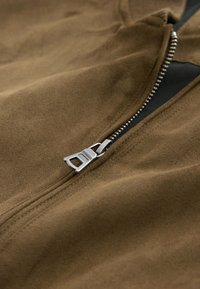 Gros plan d'une fermeture éclair argentée partiellement ouverte sur un vêtement en tissu suédé marron.