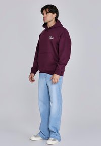 Bordeauxrode hoodie met een voorkantzak en logo, gecombineerd met lichtblauwe jeans met wijde pijpen en witte schoenen met grijze accenten.