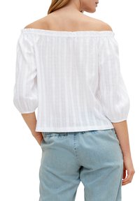 Blusa bianca off-the-shoulder con maniche a sbuffo e righe verticali, realizzata in tessuto leggero, abbinata a pantaloni in denim azzurro chiaro.