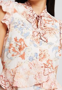 Blommig blus med en ljus krämfärgad bakgrund, prydd med rosa och blå blommor, volangärmar, krage med rosett och ett lager av volanger i fållen.