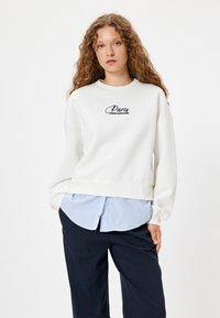 Vit sweatshirt med svart broderad "Avenue Montaigne Paris", oversized passform, ribbade ärmslut, lagd över en blårandig skjorta, mörka byxor.