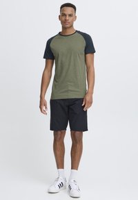 Olive green T-shirt with dark blue raglan sleeves, crew neck, cotton material, paired with black shorts and white sneakers.