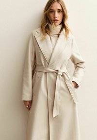 Abrigo largo beige hecho de un tejido suave, con cintura ajustada con cinturón, grandes solapas y bolsillos laterales para un diseño estructurado.