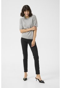 Vrouw lacht met kort donker haar, draagt een grijs gestreept shirt met korte mouwen, zwarte broek en zwarte puntige hakken tegen een witte achtergrond.