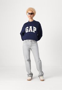 Jūrinės spalvos džemperis su balta "GAP" logotipu ir raudonu akcentu, derinamas su šviesiai pilkomis džinsomis ir baltais sportbačiais su rudos spalvos padais, vilkimas modeli.