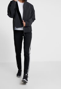 Personne portant un pantalon de survêtement noir avec des rayures blanches et des baskets noires, une veste noire partiellement zippée et une chemise blanche sur un fond uni.