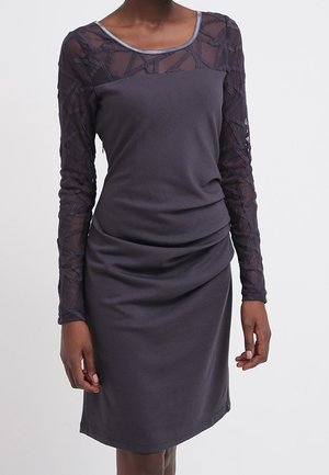 Mujer con un vestido gris oscuro hasta la rodilla, con mangas largas transparentes con estampado y un escote redondeado.
