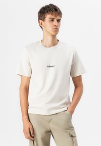 Cremefarvet bomulds t-shirt med korte ærmer, rund halsudskæring og et lille sort logo på brystet. Båret med lyse beige cargo bukser.