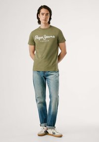 Camiseta verde oliva con el texto blanco "Pepe Jeans London". Combinada con jeans azul claro y zapatillas blancas. Tejido suave; diseño de cuello redondo.