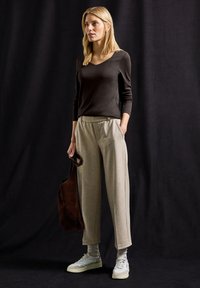 Bruna pitkähihaista puseroa, beigeä ruutuhousua, harmaita nilkkasukkia ja valkoisia tennareita. Ruskea olkutasku. Yksinkertainen musta tausta.