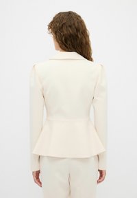 Blazer peplum crema con spalle strutturate, vestibilità su misura e dettagli della cucitura posteriore. Tessuto liscio con una leggera lucentezza.