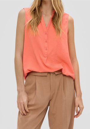 Kvinde iført ærmeløs koralfarvet bluse med knapper, som er stoppet ned i højtaljede beige bukser med folder, stående foran en ensfarvet baggrund.