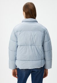 Chaqueta de plumas azul clara con cuello, que presenta un acolchado tipo baffle y puños elásticos. La parte trasera es lisa con un dobladillo recto, sin hardware visible.