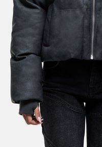 Persona che indossa una giacca imbottita nera con zip e jeans neri, con la manica piegata che mostra un foro per il pollice e una mano ben curata con unghie lunghe.