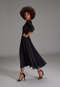 Robe de soirée noire avec un corsage pailleté, jupe en chiffon fluide, manches trois-quarts et sandales à talons hauts noires.