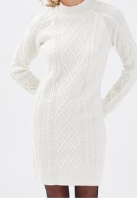 Robe blanche tricotée avec un col haut, ornée de motifs en relief et de poignets côtelés. Le design est ajusté et arrive au genou.