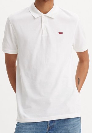 Homme portant un polo blanc à manches courtes Levi's avec des boutons, associé à un jean bleu, les mains partiellement dans les poches.