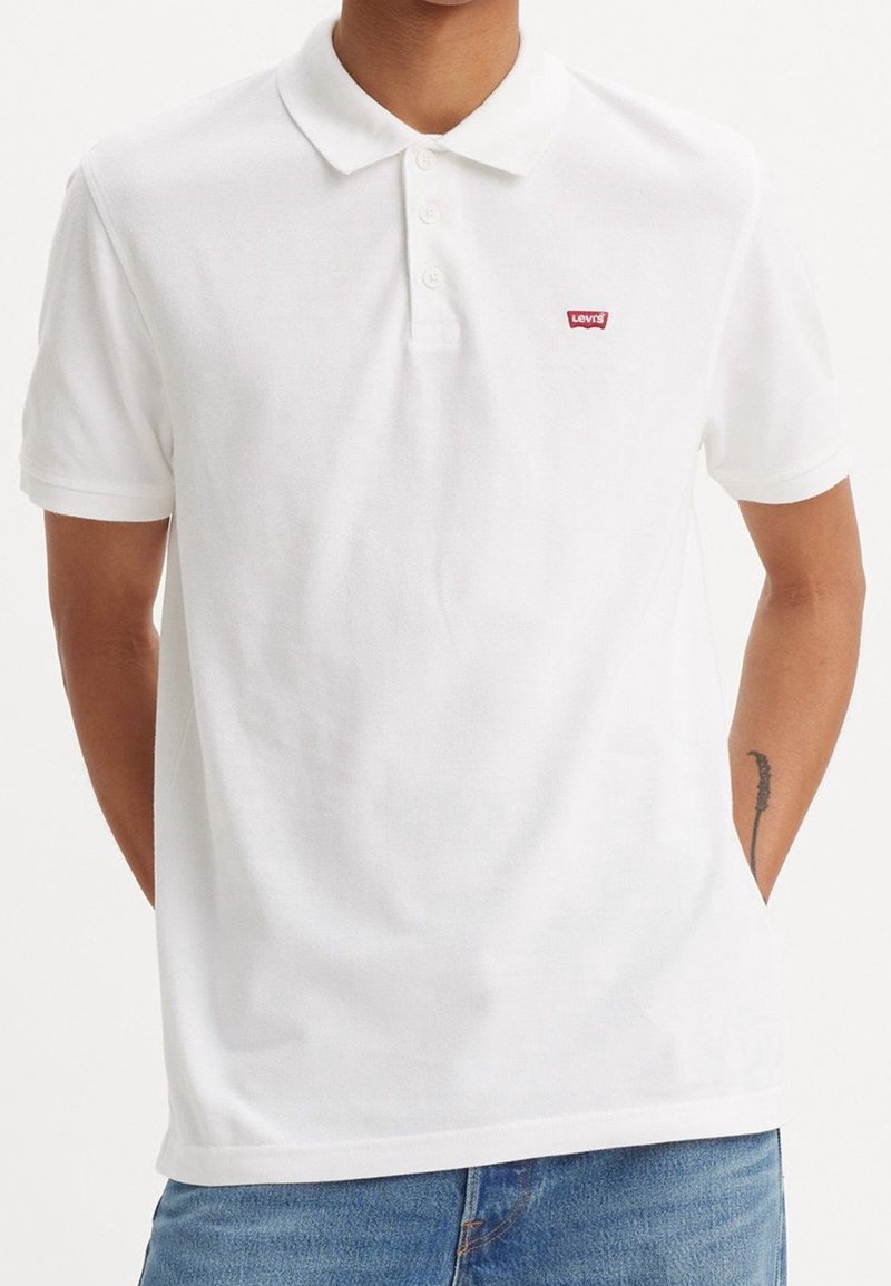 Homme portant un polo blanc à manches courtes Levi's avec des boutons, associé à un jean bleu, les mains partiellement dans les poches.