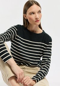 Frau in beigen Hosen und schwarzem Langarmshirt mit weißen horizontalen Streifen, im Sitzen mit übergeschlagenen Beinen, die nach links schaut.