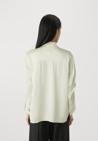 Blusa a maniche lunghe di colore giallo pallido con una trama liscia, colletto e taglio dritto. Presenta una cucitura a yoke sulla parte superiore della schiena.