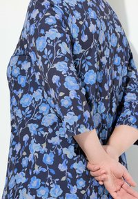 Blumenkleid mit marineblauem Hintergrund, leuchtend blauen Blumen und grünen Blättern. Drei-Viertel-Ärmel und eine lockere Passform sind sichtbar.