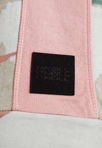 Étiquette carrée en tissu noir avec le texte répété "O'NEILL" cousue sur un tissu rose avec détail de couture.