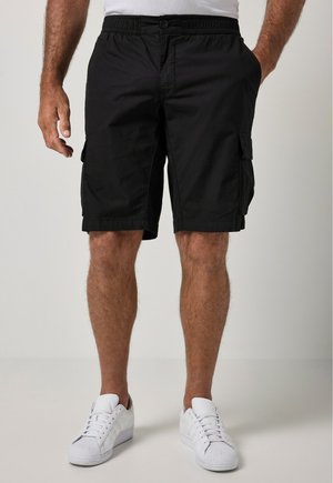 Mand iført sorte cargoshorts, hvide sneakers og en hvid skjorte, stående med den ene hånd i lommen og den anden afslappet ved siden.