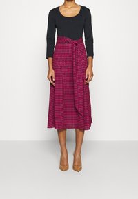 Svart långärmad topp i kombination med en knälång rosa kjol med houndstooth-mönster, knuten vid midjan med ett matchande bälte.