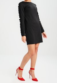 Femme portant une robe noire à manches longues avec des manches volantes et des chaussures à talons hauts rouges avec bride de cheville, se tenant devant un fond blanc.