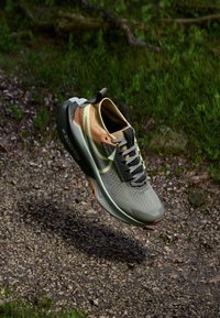 Chaussure de course avec une tige en mesh gris et beige, accents verts, texture perforée, et une semelle noire avec un logo orange vif.