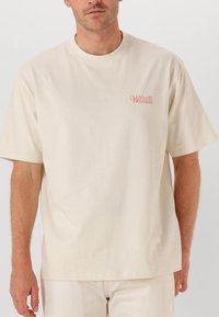 Beige bomullströja med kort ärm och rundad halsringning. Litet logga i korall på vänster bröstområde. Slät textur, avslappnad passform.