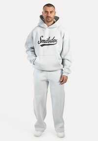 Hombre de pie con sudadera ligera gris y pantalones de chándal a juego con el logo "Smilodox", combinados con zapatillas blancas sobre un fondo liso.