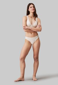 Ensemble de lingerie beige comprenant un bralette texturé avec un décolleté en V profond et un string assorti avec une taille logo. Tissu doux et design ajusté.