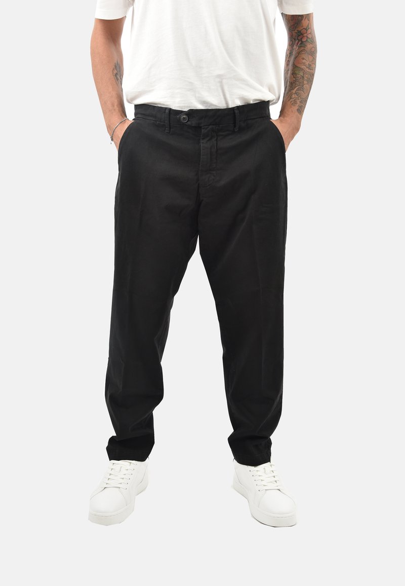 Persona con pantaloni casual neri, sneakers bianchi e una maglietta bianca, con le braccia tatuate visibili e le mani in tasca, su sfondo bianco.