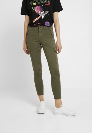 Person iført olivengrønne cargobukser, hvide sneakers og en sort t-shirt med farverige rumtema-grafikker og tekst.