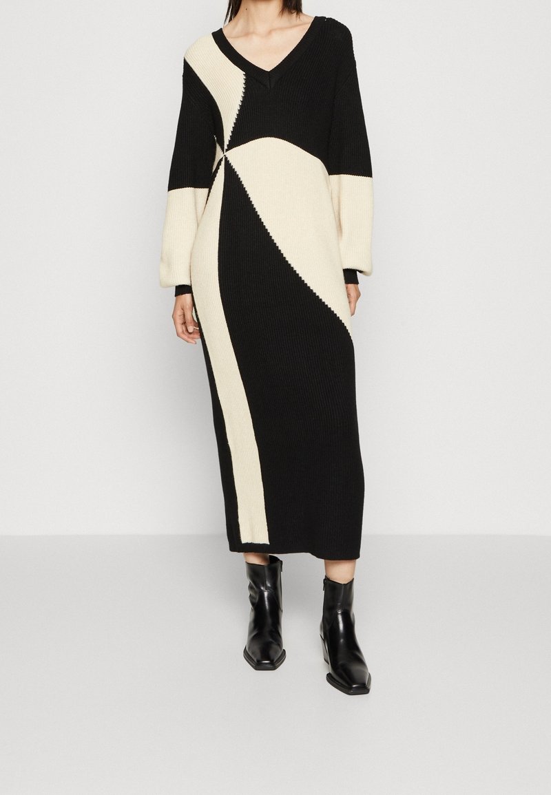 Robe en tricot noir et crème avec un décolleté en V et des manches longues, présentant un design géométrique en blocs de couleurs et une texture côtelée. Portée avec des bottines noires.