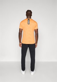 T-shirt de manga curta com padrão de camuflagem laranja, com um rótulo preto apresentando a palavra "BORG" nas costas, combinado com calças pretas e ténis brancos.