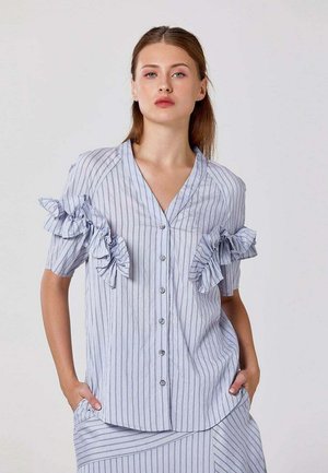 Femme portant un chemisier bleu clair à rayures verticales avec boutons sur le devant et détails de volants aux manches, associé à une jupe rayée assortie.