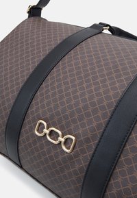 Sac de voyage en tissu à motif marron avec des accents en cuir noir et une sangle ajustable. Présente un détail en chaîne dorée à l'avant.