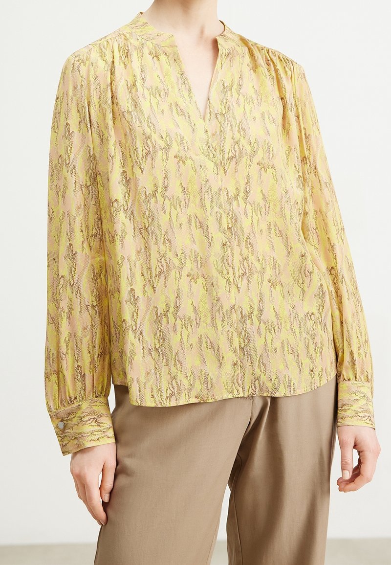 Blusa a maniche lunghe di un giallo chiaro con un motivo testurizzato di verdi e rosa tenui. Presenta un collo a V e maniche con polsini.