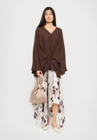 Woman with long dark hair wearing a brown oversized top and floral asymmetrical skirt, holding a beige satin handbag with white heels.