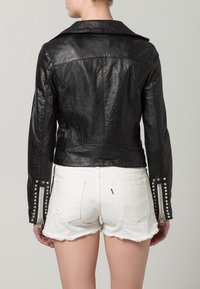 Frau, die eine schwarze Lederjacke mit silbernen Nieten und Reißverschlüssen an den Ärmeln trägt, kombiniert mit weißen ausgefransten Denim-Shorts, von hinten gezeigt.