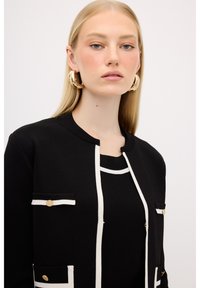 Svart tröja med vita accenter och fickdetaljer, med rund halsringning och textilstruktur. Modellen bär stora, runda örhängen.