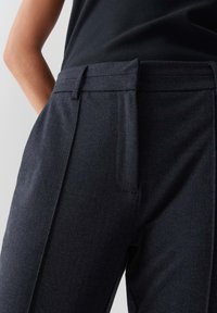 Close-up of person wearing dark tailored trousers and a black top, showing waistband, belt loops, and front pleat details.