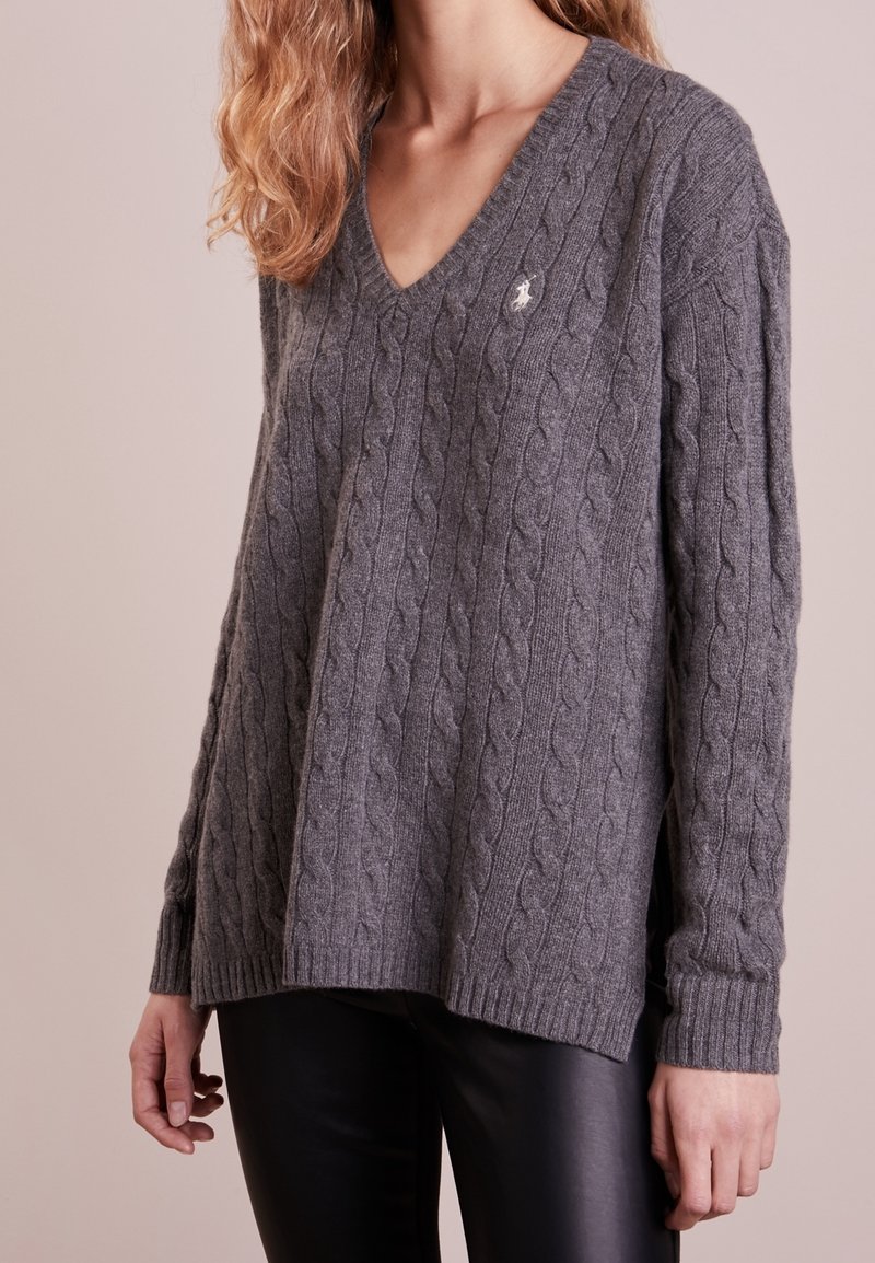 Femme portant un pull gris à col en V en maille torsadée, manches longues, et un pantalon noir, debout devant un fond uni.