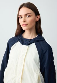 Gevoerde jas met een marineblauwe bovenkant en een crème-kleurige onderpaneel. Lichte stof met een licht gekreukt textuur en een ritssluiting aan de voorkant.
